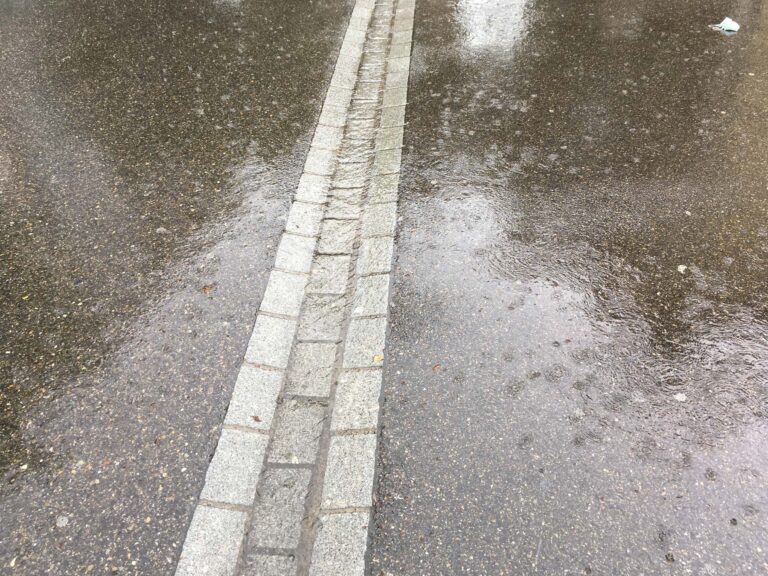 Dienstleistung Siedlungs- und Umgebungsentwässerung: Terraingestaltung zur Ableitung von Regenwasser. Steine eingelassen im Asphalt bilden eine Abwasserrinne.